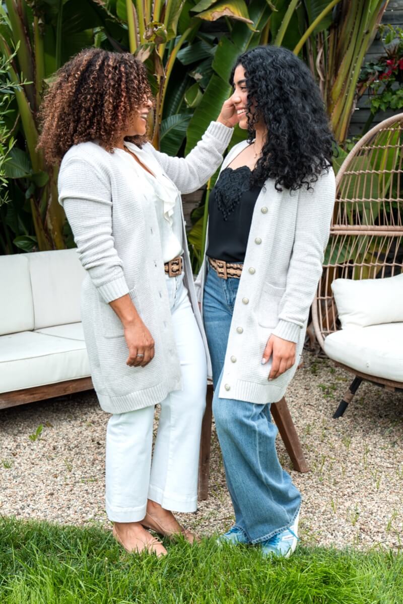 Mom model wearing the Cozy Cardigan in Dove,  tread belt in multi, Showstopper Top in White, and the Palm Beach Crop in White. Daughter model wearing the Cozy Cardigan in Dove,  tread belt in multi, Ashbury Jean in Faded Wash, Baroque Cami in Black