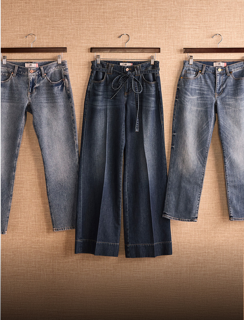 3 pairs of denim positioned on coat hangers, hung against a linen wall.